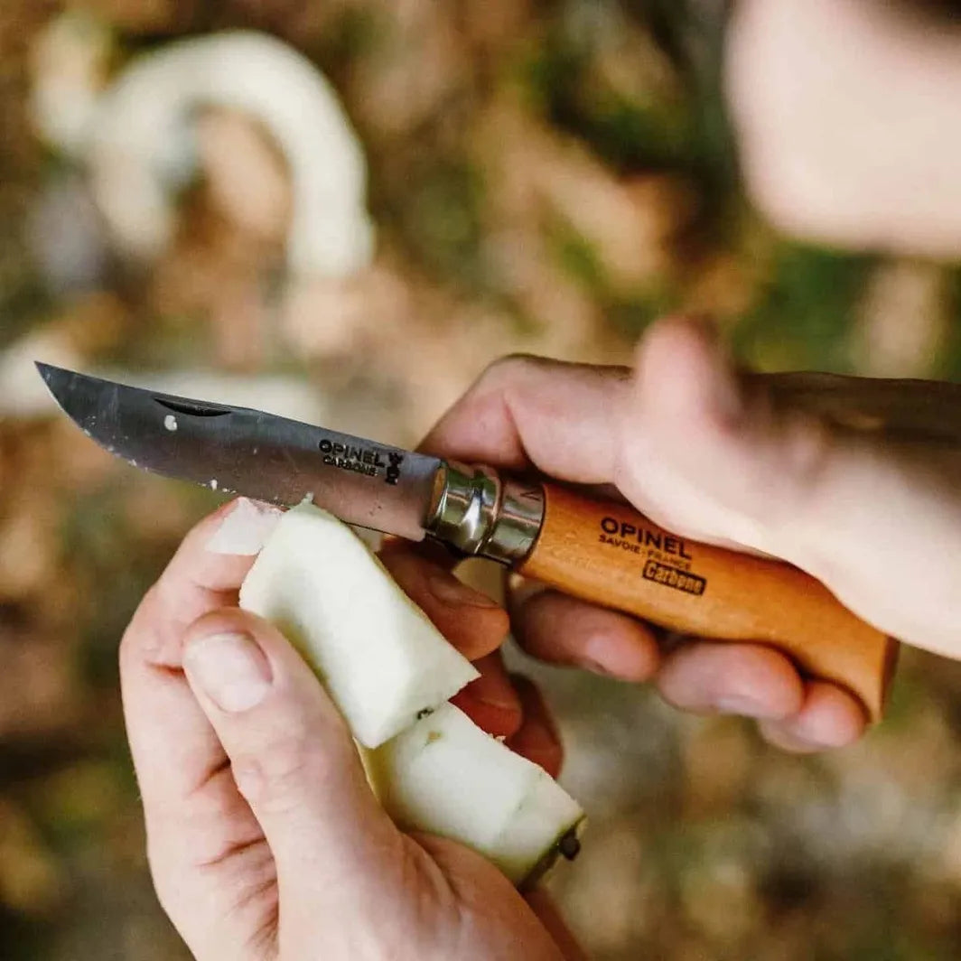 Person using an Opinel #8 carbon folding knife to cut a piece of fruit with a blurred natural background brought to you by Homestead Iron