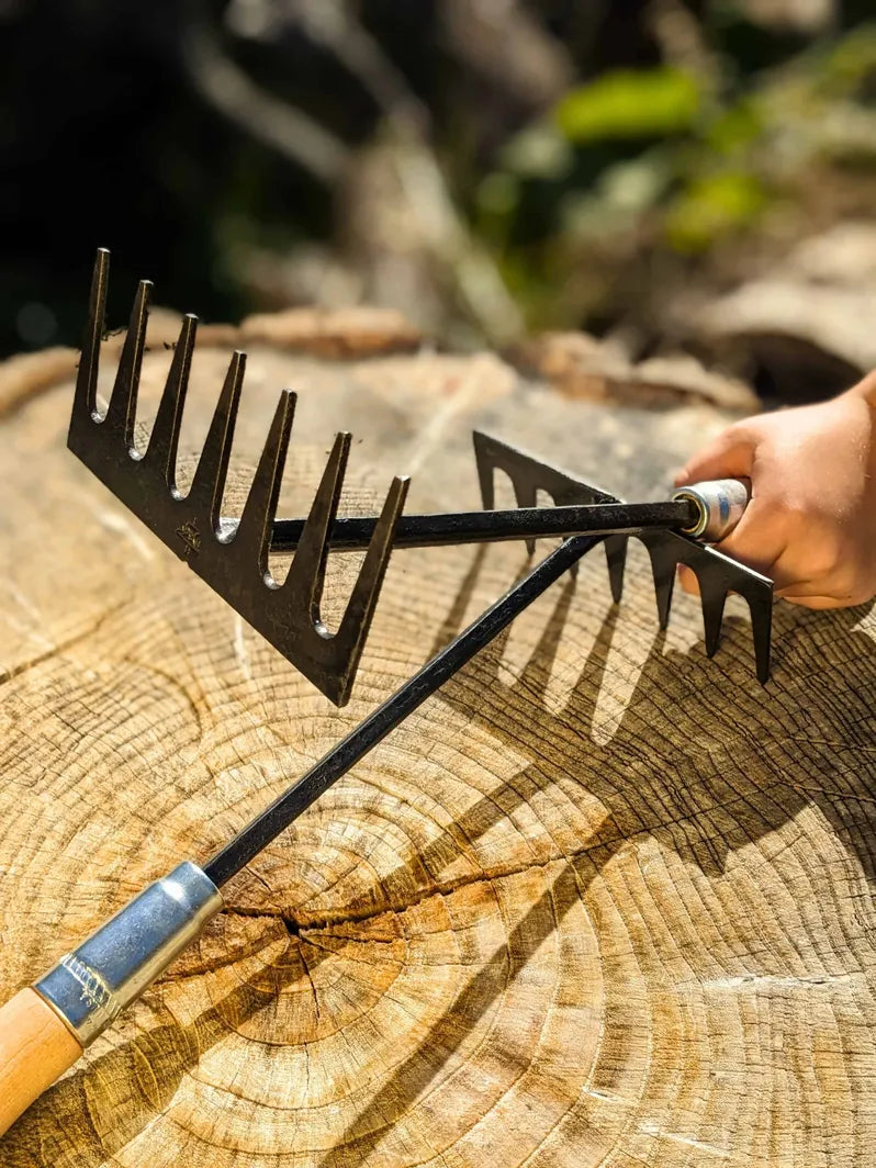 Hand Rake hand forged by Homestead Iron held by a hand on wooden background with deep shadows. 