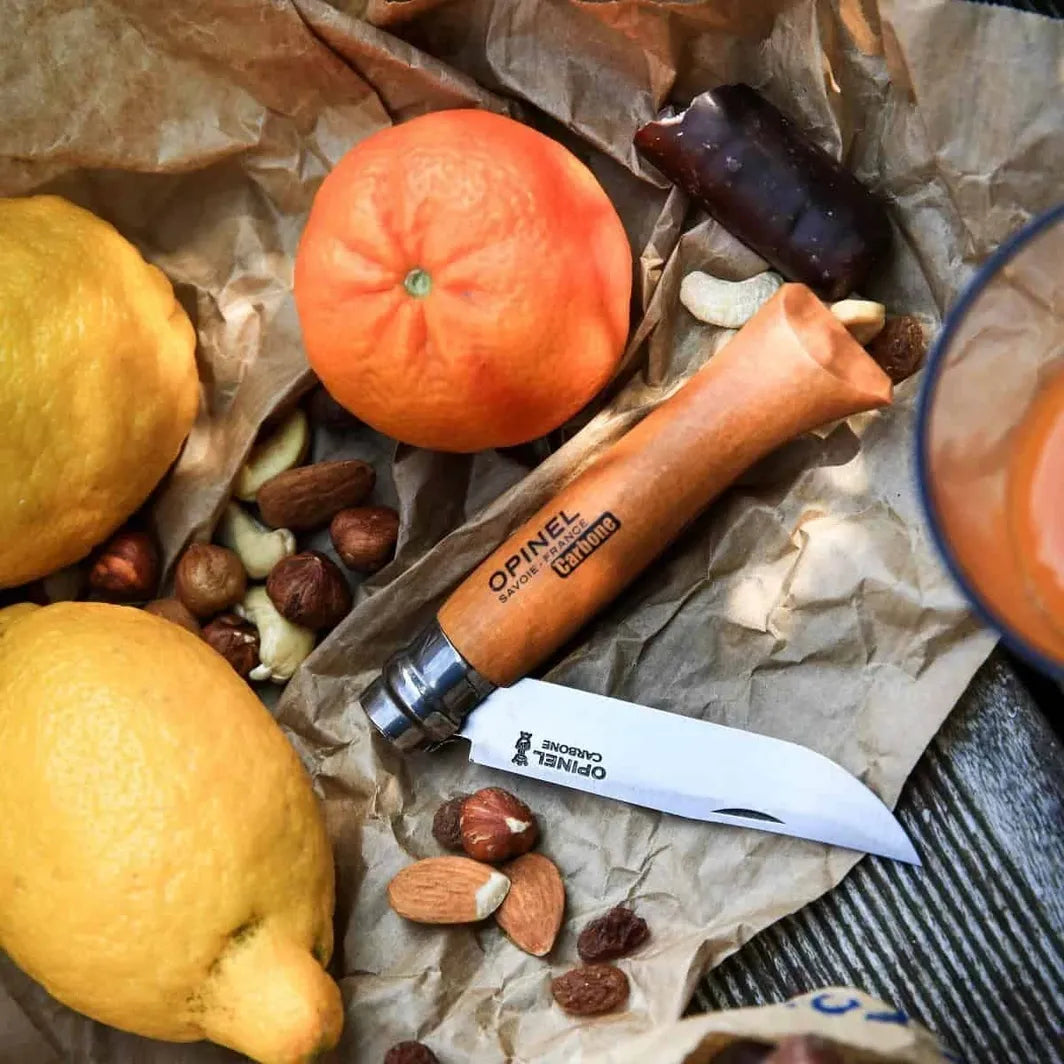 Opinel #8 folding carbon knife on a wooden surface with lemons, oranges, and nuts.